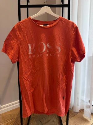 Orange t-shirt från Hugo Boss - Snygg orange t-shirt från Hugo Boss med stort vitt BOSS-tryck framtill. Klassisk rund halsringning och korta ärmar. Tillverkad i mjuk bomull som känns skön mot huden. Perfekt för dig som vill sticka ut med en färgstark look.