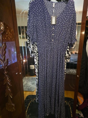 Blå långklänning med vita blommor Only Carmakoma - Supersöt långklänning från Only Carmakoma i mörkblått med små vita blommor. Klänningen har v-ringning, knappar framtill, korta ärmar med volangdetalj och ett lätt, luftigt tyg. Perfekt för dig som gillar en flowy och feminin stil.