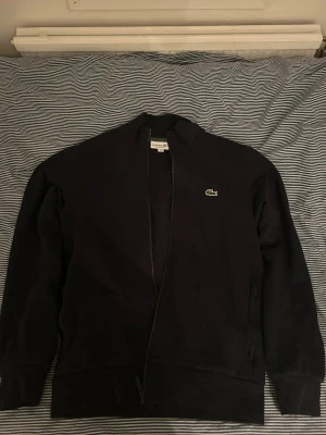 Mörk blå full zip tröja från Lacoste - Blå full zip tröja från Lacoste i storlek S med klassisk krokodillogga på bröstet. Regular fit, storlek S.Tröjan har använts en gång, nytt skick 10/10! Nypris: 1599kr. 