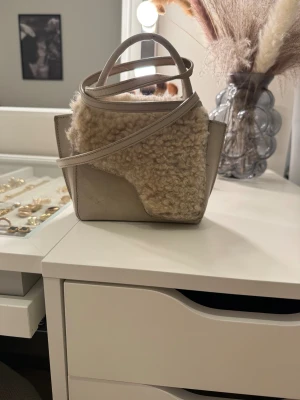 Beige handväska med pälsdetaljer - Snygg beige handväska med både handtag och axelrem. Väskan har en unik design med mjuka pälsdetaljer och slät skinn på sidorna. Perfekt storlek för att bära med sig det viktigaste och ger en cool, trendig vibe till din outfit.