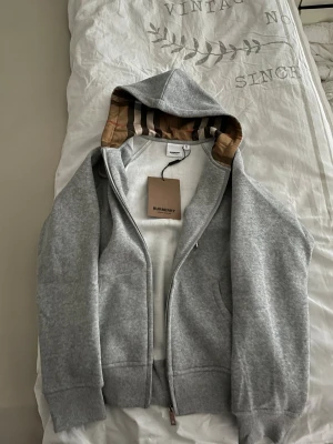 Grå Burberry hoodie med logga, M - Snygg grå hoodie från Burberry i storlek M. Tröjan har huva med klassiskt Burberry-randigt foder, dragkedja framtill och snörning vid halsen. Stor svart Burberry-logga på bröstet och mjukt bomullsmaterial. Perfekt för dig som gillar stilrena märkesplagg.