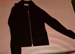 Svart kofta med dragkedja från Norrie - Svart, stilren kofta från Norrie med hel dragkedja framtill. Tröjan har långärmad design och en enkel, clean look som passar perfekt till jeans eller kjol. Materialet känns mjukt och är troligtvis stickat. Snygg att ha öppen eller stängd.