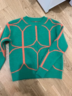 Grön-orange stickad tröja med mönster - Cool stickad tröja i grönt med grafiskt orange mönster. Tröjan har rund halsringning, ribbade muddar och är gjord i ett mjukt material som känns skönt mot huden. Perfekt för dig som vill sticka ut med färg och stil.