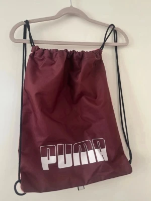 Vinröd gympåse från Puma - Snygg vinröd gympåse från Puma med stort vitt logotryck framtill. Väskan har svarta snören som axelband och stängs med dragsko. Perfekt för träning eller när du vill bära med dig det viktigaste. Tillverkad i slitstarkt tygmaterial.