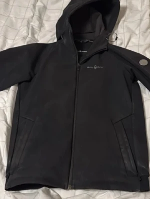 Svart vindjacka från Sail Racing - Svart vindjacka från Sail Racing med huva och dragkedja framtill. Bra skick, lite skav på ärmarna.