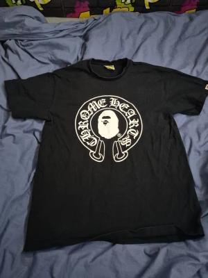 Svart Chrome Hearts x BAPE T-shirt - Säljer en svart T-shirt från Chrome Hearts och BAPE med stor vit logga och grafik på både fram- och baksidan. Framsidan har Chrome Hearts-tryck med BAPE-ansikte, baksidan har BABY MILO och Chrome Hearts-kors med BAPE-figur. Klassisk passform och rund hals.