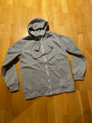 Grå hoodie från Polo Ralph Lauren - Snygg grå hoodie från Polo Ralph Lauren med dragkedja framtill och klassisk huva med dragsko. Broderad mörkblå logga på bröstet och två fickor fram. Tillverkad i mjuk bomullsblandning som känns skön mot huden. Perfekt för en avslappnad och stilren look.