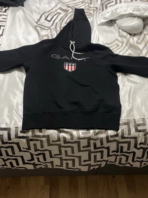 Svart hoodie från GANT med logga - Svart hoodie från GANT med vit dragsko i huvan och stor GANT-logga samt ett rött, vitt och blått sköldmärke på bröstet. Klassisk känguruficka framtill och ribbade muddar. Tillverkad i mjukt bomullsmaterial för en skön känsla.