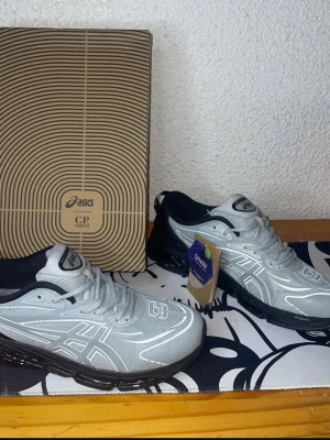 Asics x CP Company grå sneakers - Snygga Asics x CP Company sneakers i grått med vita detaljer och svart sula. Modellen är GEL-Quantum 360 och har Ortholite-innersula för extra komfort. Skorna har en sportig design med reflekterande detaljer och tydlig logga på tungan och sidan.