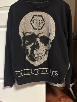 Svart långärmad topp Philipp Plein - Cool svart långärmad topp från Philipp Plein med stor döskallegrafik framtill och logga både på bröstet och vid fållen. Rund halsringning och mjukt material som känns skönt mot huden. Perfekt för dig som gillar edgy streetstyle.