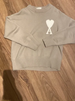 Beige stickad tröja Ami Paris - Snygg beige stickad tröja från Ami Paris med deras ikoniska vita hjärta och A-logga på bröstet. Tröjan har rund hals, ribbade muddar och nederkant. Perfekt för dig som gillar stilrena och trendiga plagg.