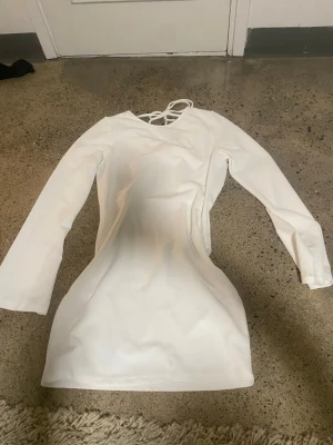 Vit långärmad miniklänning - Trendig vit klänning  med långa ärmar och figurnära passform. Klänningen har en snygg drapering framtill och dekorativa knytband i ryggen. Perfekt för dig som gillar stilrena och moderna plagg. 