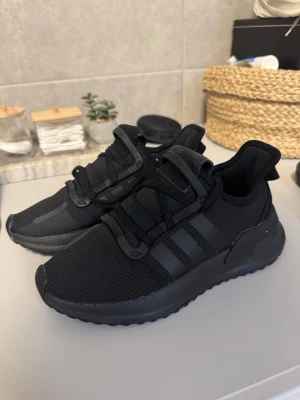  Adidas sneakers - Snygga helsvarta sneakers från Adidas med klassiska tre ränder på sidan. Ovandelen är i ventilerande mesh och detaljer i syntet. Skorna har en chunky sula och modern design som passar perfekt till streetwear.