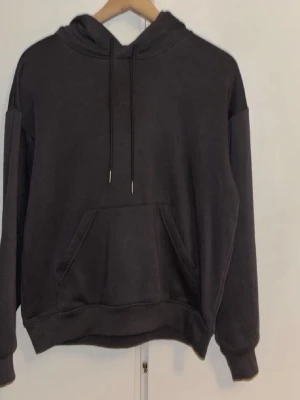 Svart hoodie med ficka och huva - En basic svart hoodie med stor magficka och justerbar huva med snören. Hoodien har ribbade muddar vid ärmar och nederkant, samt en avslappnad passform. Perfekt för en chill och enkel stil.