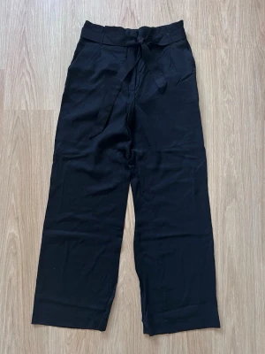 Svarta kostymbyxor med knytband i midjan från H&M, storlek 40 - Stilrena svarta kostymbyxor med hög midja och knytskärp. Raka, vida ben och sidofickor. Nyskick, knappt använda!