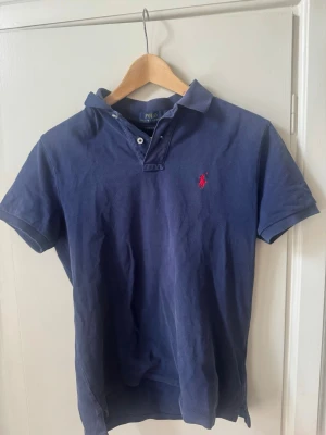 Marinblå polo piké från Ralph Lauren - Klassisk marinblå pike från Ralph Lauren med röd broderad logga på bröstet. Slim fit-modell i mjuk bomull med korta ärmar och krage med knappar. Passar om du är cirka 180 cm långPerfekt för dig som gillar stilrena och tidlösa plagg. Nypris 1700
