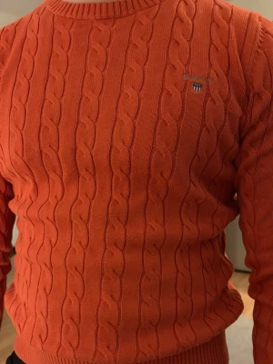 Orange kabelstickad tröja från Gant - Säljer en orange kabelstickad tröja från Gant med rund halsringning och broderad logotyp på bröstet. Tröjan har ribbade muddar och är perfekt för dig som gillar klassisk preppy stil.