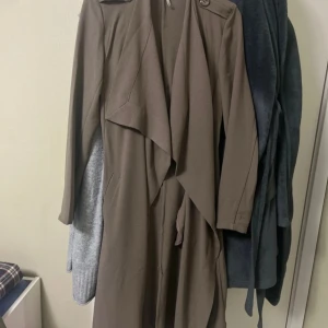 Object kappa / trenchcoat - Brun trenchcoat i lång modell med knyskärp / bälte i midjan och axelklaffar med knapp. Jackan har en klassisk omlott-design och faller snyggt framtill. Aldrig använd