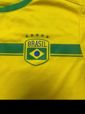 Gul Brasil fotbollströja - Snygg gul Brasilien fotbollströja med gröna detaljer och tryckt BRASIL-emblem på bröstet. Tröjan har rund halsringning med grön kant och korta ärmar med gröna muddar. Perfekt för match eller träning. Tror det är 170 cm passar ungefär 14-16 åringar 