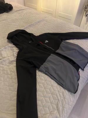 Svart och grå vindjacka The North Face - Snygg vindjacka från The North Face i svart och grått med huva och dragkedja. Jackan har en bröstficka med dragkedja och logga på bröstet. Perfekt för blåsiga dagar och outdoor-aktiviteter. Materialet är lätt och smidigt. En bit av dragkedjan är av så det går ej att knäppa jackan, men det går att fixa. Men annars är jackan i väldigt bra skick.