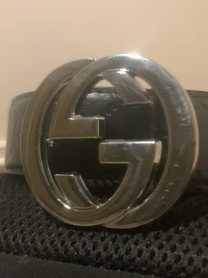Svart Gucci bälte med GG-spänne - Säljer ett svart bälte från Gucci med det klassiska dubbla GG-spännet i silver. Bältet är gjort i präglat skinn med GG-loggor längs hela remmen. Perfekt accessoar för att lyfta vilken outfit som helst.