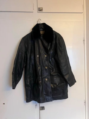 Svart lång skinnjacka med pälskrage - Säljer en svart lång skinnjacka med dubbelknäppning och bred pälskrage. Jackan har stora fickor framtill och en klassisk rak passform. Perfekt för dig som vill ha en tidlös och cool look. Pälskragen fästs med knappar (saknas en knapp)