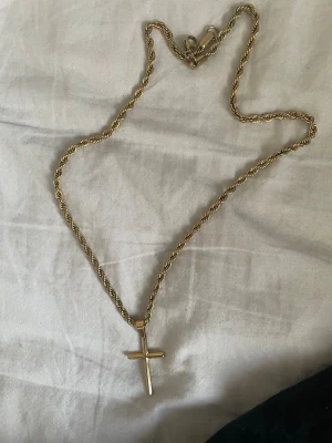 Guldhalsband med korshänge - Snyggt halsband i guldfärgad metall med tvinnad kedja och ett korsformat hänge. Kedjan har en klassisk karbinhake och hänget är stilrent och enkelt, vilket gör det lätt att matcha med olika outfits. Perfekt accessoar för dig som gillar minimalistisk design.