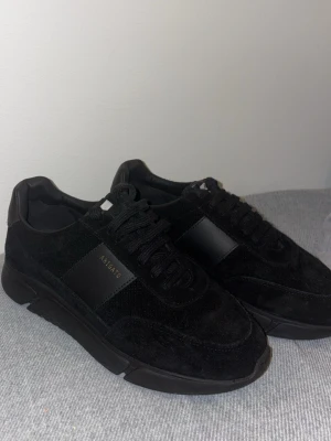 Svarta sneakers från Axel Arigato - Snygga helsvarta sneakers från Axel Arigato med ovandel i mocka och detaljer i skinn. Klassisk siluett med snörning och diskret logga på sidan. Perfekta för dig som gillar stilrena och tidlösa skor.