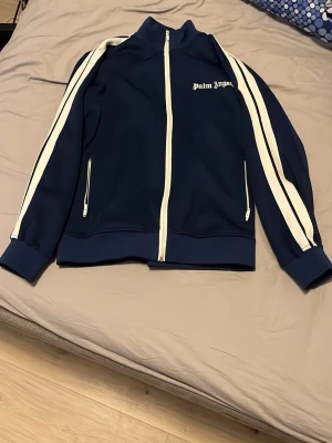 Blå zip-tröja från Palm Angels - Snygg marinblå zip-tröja från Palm Angels med vita ränder längs ärmarna och vit dragkedja. Tröjan har hög krage, två fickor med dragkedja och logga på bröstet. Perfekt för en sportig och trendig look.