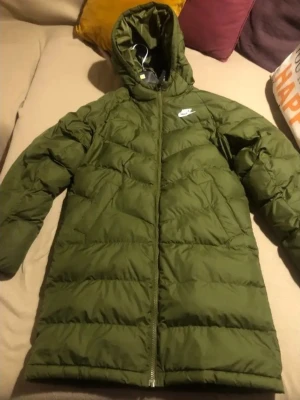 Grön dunjacka med huva från Nike - Snygg grön dunjacka från Nike med huva och dragkedja framtill. Jackan har en quiltad design och Nike-logga på bröstet. Perfekt för kalla dagar och ger en sportig look. Passar dig som vill ha både stil och värme under vintern.