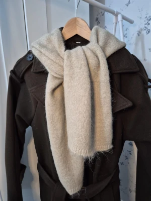 Balaklava  - Säljer en beige stickad väst med huva. Västen är i mjukt och fluffigt material och har en enkel, rak passform utan knappar eller dragkedja. Perfekt att slänga över en tröja för extra värme. Stilren och enkel design som passar till många outfits. 