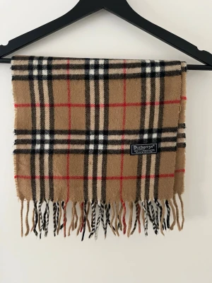 Halsduk från Burberry - Snygg halsduk i ull från Burberry med det ikoniska beige, svarta, vita och röda rutmönstret. Halsduken har fransar i ändarna och en vävd logotypetikett. Perfekt accessoar för att lyfta din outfit.