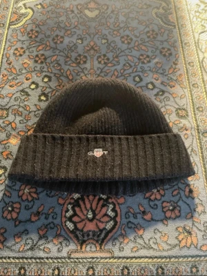 Svart ribbstickad mössa från GANT - Svart ribbstickad mössa från GANT med uppvikt kant och broderad logotyp framtill. Klassisk och enkel design som passar perfekt till kalla dagar. Tillverkad i mjukt material för extra komfort.