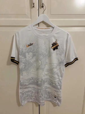 Vit AIK fotbollströja från Nike - Snygg vit AIK fotbollströja från Nike med guldfärgade detaljer och klubbmärke på bröstet. Tröjan har korta ärmar med svarta och gula ränder vid ärmsluten samt ett unikt mönster framtill. Perfekt för dig som älskar fotboll och AIK.