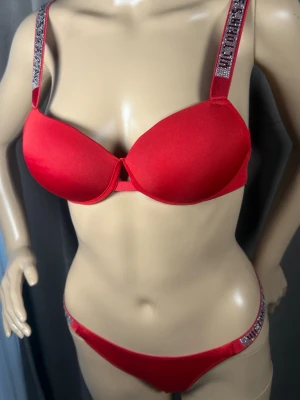 Rött bh- och trosa-set med strass - Snyggt rött underklädesset med vadderad bygel-bh och matchande trosa. Axelbanden och sidorna på trosan är dekorerade med glittrande strassdetaljer. Setet har en modern och ungdomlig design, perfekt för dig som gillar att sticka ut.