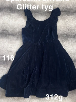Mörkblå sammetsklänning med volang - Mörkblå klänning i sammetsmaterial med glittrigt tyg och volangdetaljer vid axlarna. Söt rosett i midjan och ärmlös design. Perfekt för dig som vill sticka ut lite extra.🌸 Sparsamt använd. Glittertyg. Storlek 116