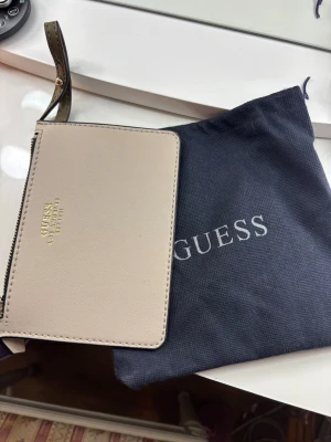 Beige plånbok från Guess - Snygg beige plånbok från Guess med guldfärgad logga framtill. Plånboken har dragkedja och går att fästa på väskan. Tillverkad i slätt konstläder och levereras med en mörkblå dustbag med Guess-logga.