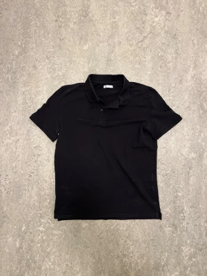 Svart piké/polo COS - Klassisk svart pikétröja från Cos med krage och knappar framtill. Tröjan har korta ärmar och är tillverkad i ett mjukt bomullsmaterial som känns skönt mot huden. Enkel och stilren design som passar till det mesta.