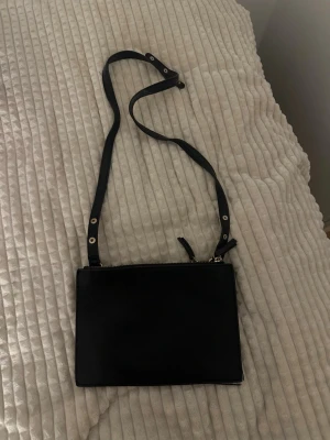 Svart axelväska i skinnimitation - Snygg svart axelväska med justerbart axelband och silverfärgade detaljer. Väskan har en stilren, rektangulär form och stängs med dragkedja upptill. Perfekt för dig som gillar minimalistisk och clean stil.