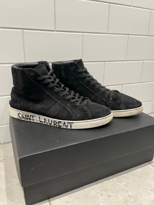 Svarta Saint Laurent high top sneakers - Svarta high top sneakers från Saint Laurent i mocka med vita sulor och handskriven logga på sidan. Klassisk siluett med svarta skosnören och diskreta sömmar. Perfekta för en stilren och edgy look. Dem e i bra skick enda är en liten defekt på undersidan men inget man märker.