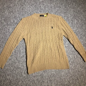 Beige kabelstickad tröja Polo Ralph Lauren S-M - Snygg beige kabelstickad tröja från Polo Ralph Lauren med rund halsringning och den klassiska broderade loggan på bröstet. Tröjan har ribbade muddar vid ärmslut och nederkant. Perfekt för lager på lager och en stilren vardagslook. Kontakta vid frågor om pris och storlek 😊