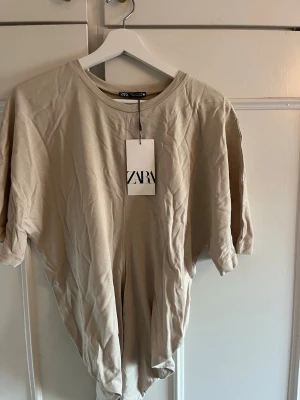 Beige t-shirt från Zara - Snygg beige t-shirt från Zara i mjuk bomull. Klassisk rund halsringning och korta ärmar. Enkel och stilren design som passar till det mesta. Perfekt basplagg för garderoben.Ny med prislapp på! 