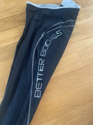 Svarta Better Bodies tights med dragkedja - Snygga svarta tights från Better Bodies med silverfärgade sömmar och logga längs benet. De har dragkedja vid benslut och är tillverkade i ett stretchigt syntetmaterial som passar perfekt till träning. Midjan är normal och insidan är ljusgrå. 3/4 längd. 