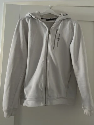 Vit hoodie från Sail Racing - Vit hoodie med dragkedja från Sail Racing. Har en bröstficka med dragkedja och logga på vänster sida. Klassisk huva och långa ärmar. Perfekt för en chill och sportig stil.