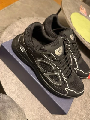 Svarta Dior B30 sneakers - Snygga svarta Dior B30 sneakers med vita detaljer och reflekterande linjer. Skorna har en sportig siluett, mesh och läderpaneler samt tydlig Dior-logga på plösen och sidan. Perfekta för dig som vill ha en exklusiv och trendig look.