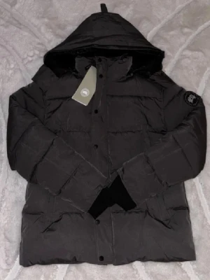 Svart dunjacka från Canada Goose - Snygg svart dunjacka från Canada Goose med huva och diskret logotyp på ärmen. Jackan har en klassisk puffer-design och är tillverkad i slitstarkt material som håller värmen under kalla vinterdagar. Perfekt för dig som vill ha både stil och funktion.