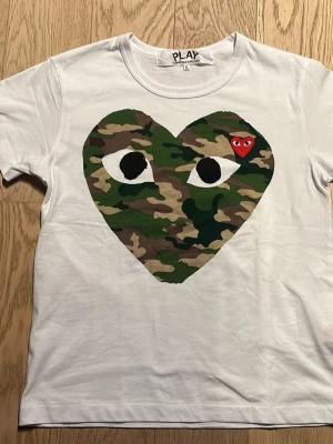 Vit Comme des Garçons PLAY t-shirt - Säljer en vit t-shirt från Comme des Garçons PLAY med det ikoniska hjärtat i camouflage och ett litet rött hjärta på bröstet. T-shirten har rund hals och korta ärmar. Perfekt statement-plagg för dig som gillar streetwear och unika prints. Pris kan diskuteras