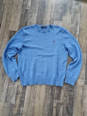 Blå stickad tröja från Polo Ralph Lauren - Klassisk blå stickad tröja från Polo Ralph Lauren med rund halsringning och ribbade muddar. Tröjan har det ikoniska lilla röda Polo-logot på bröstet och är tillverkad i mjukt material. Perfekt för lager på lager och enkel att matcha.