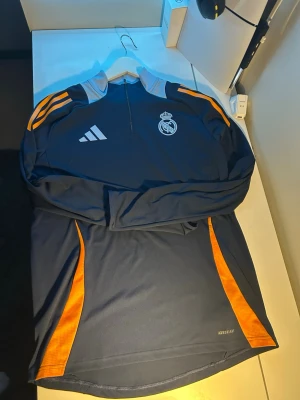 Real Madrid träningströja Adidas - Mörkblå långärmad träningströja från Adidas med Real Madrid-logga på bröstet. Tröjan har orange detaljer längs sidorna och axlarna, halv dragkedja och står 'AEROREADY' på nederkanten. Perfekt för fotbollsträning. Den är L men passar ochså som en M bra