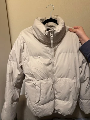 Beige pufferjacka med hög krage - Säljer en stilren beige pufferjacka med hög krage och dragkedja framtill. Jackan har två snedställda fickor och quiltad design som ger en schysst volym. Perfekt för kalla vinterdagar när du vill hålla värmen och se snygg ut.